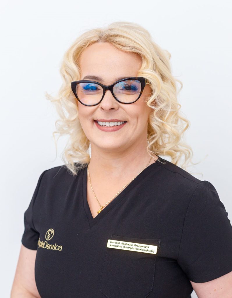 Portret uśmiechniętej kobiety o blond, kręconych włosach, ubranej w czarny uniform medyczny z logo „ImplaDentica”. Kobieta nosi okulary w ciemnych oprawkach typu „kocie oczy” i złoty łańcuszek.