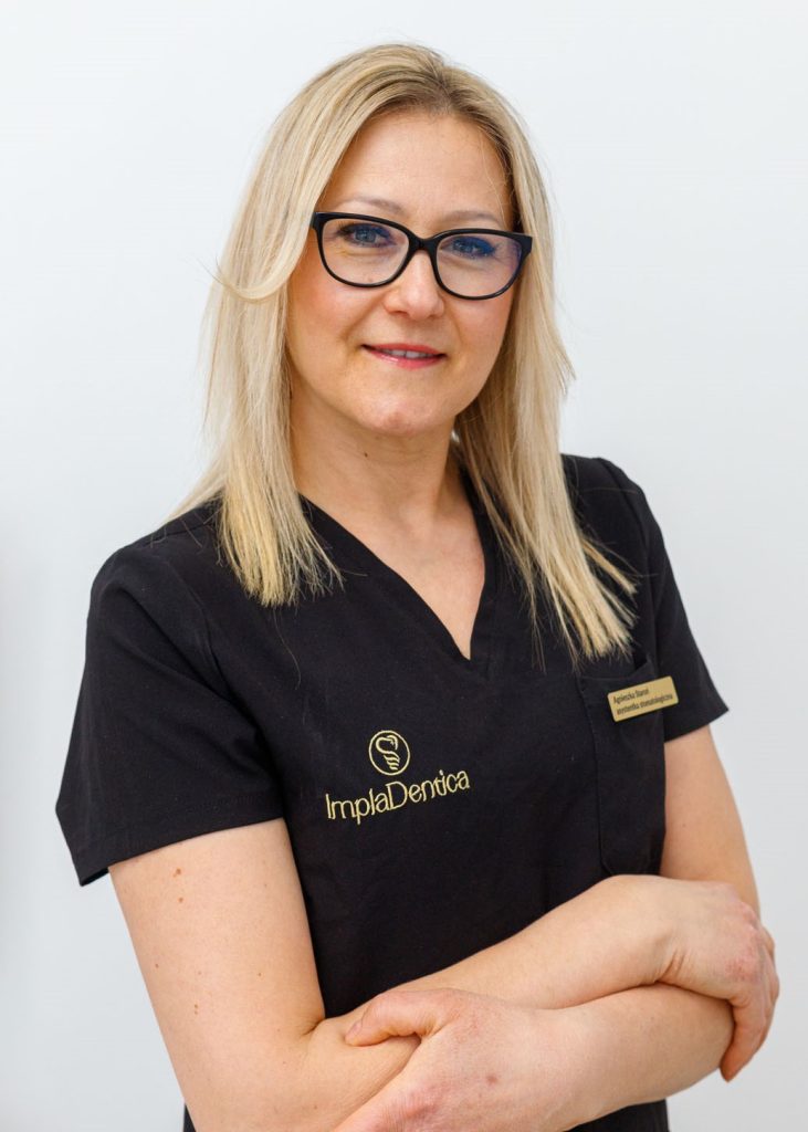 Portret uśmiechniętej kobiety w czarnej bluzce medycznej z logo „ImplaDentica” i identyfikatorem. Kobieta ma blond włosy, nosi czarne okulary i ma splecione ręce na piersiach.