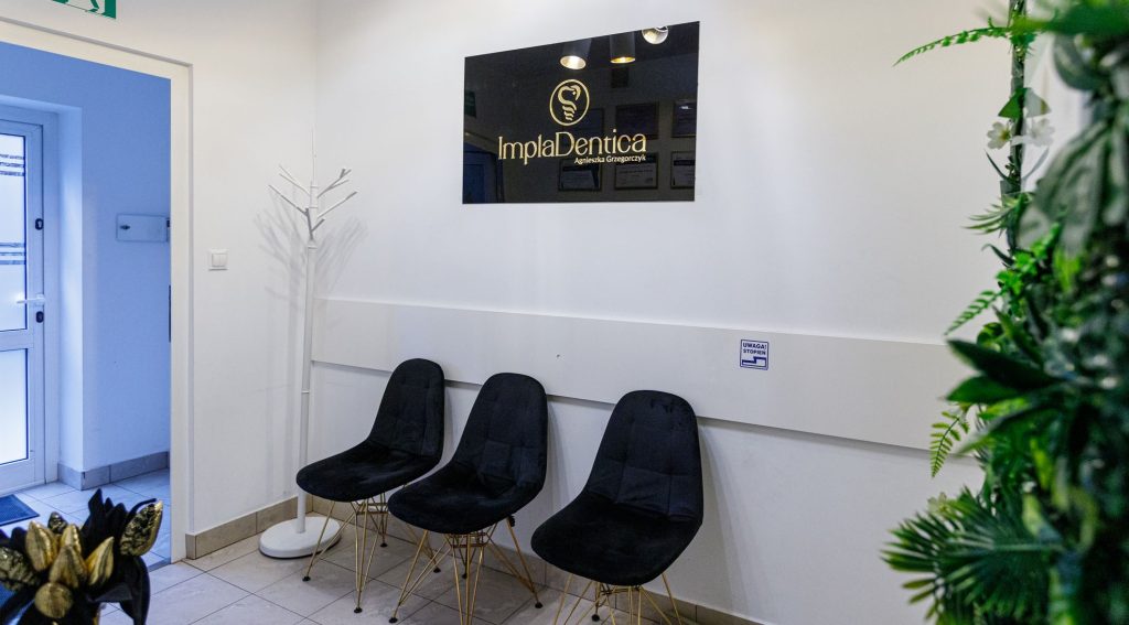 Wnętrze nowoczesnej poczekalni stomatologicznej ImplaDentica. Na białej ścianie wisi czarny błyszczący szyld z logotypem firmy, a pod nim stoją trzy eleganckie, czarne krzesła na drewnianych nogach. Po prawej stronie widoczny jest fragment zielonej ściany z roślin.
