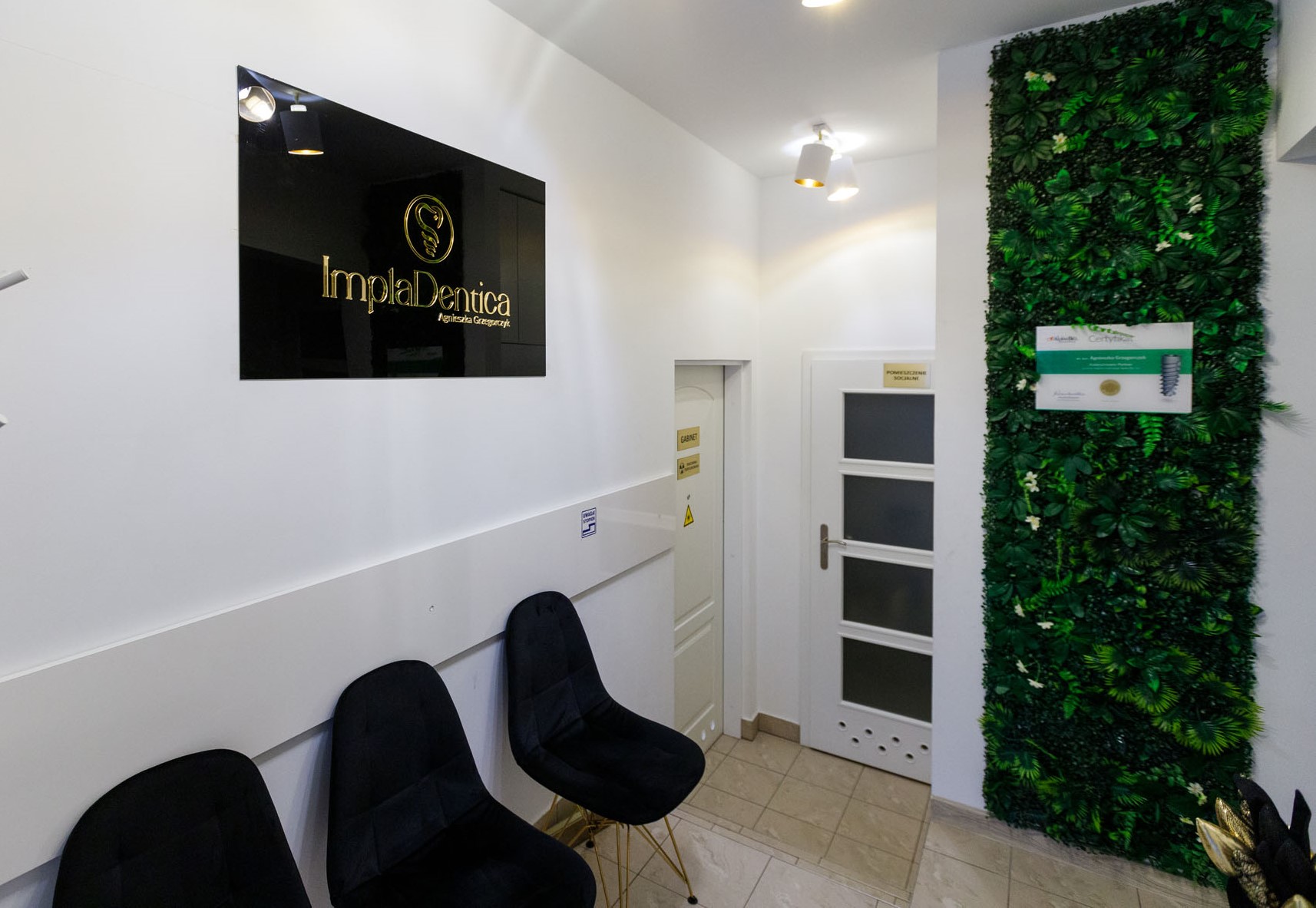 Perspektywa korytarza w klinice stomatologicznej. Na ścianie widnieje czarny szyld z logo firmy, obok znajdują się białe drzwi oraz dekoracyjna ściana z zielonej roślinności. W dolnej części widać nowoczesne krzesła dla pacjentów.