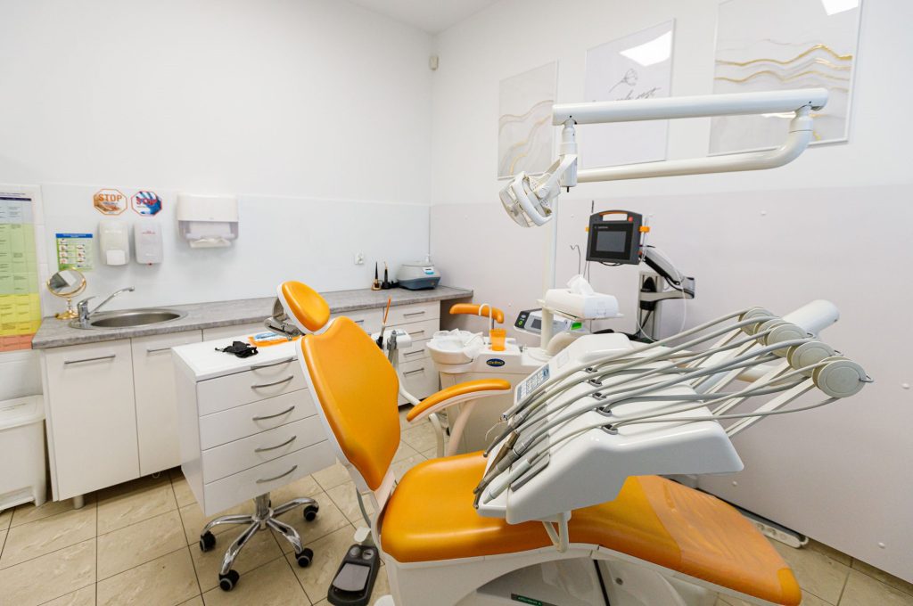 Nowoczesny gabinet stomatologiczny z pomarańczowym fotelem dentystycznym w centrum. Po lewej stronie znajdują się białe szafki z umywalką i dozownikami, a w tle specjalistyczny sprzęt medyczny oraz obrazy na ścianie.