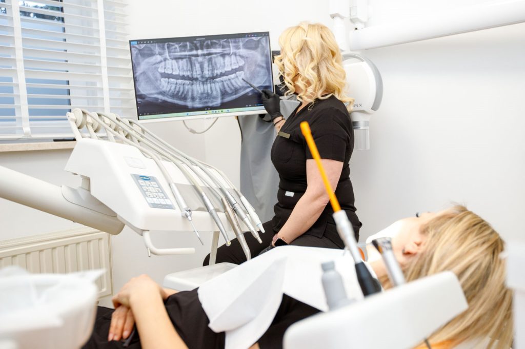 Specjalistka w czarnym stroju medycznym i rękawiczkach pokazuje pacjentce zdjęcie rentgenowskie szczęki na monitorze. Pacjentka leży na fotelu dentystycznym, widoczna z profilu w dolnej części kadru.